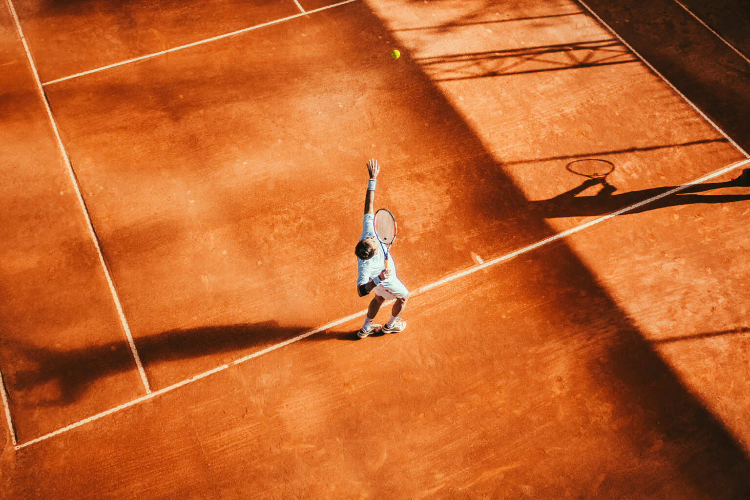Aerial view of a tennis player serving on a clay court in late-afternoon light